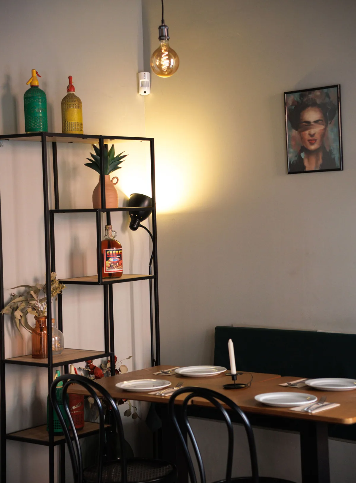 Table laid for two beside open shelving with bottles and a plant, under a filament pendant
