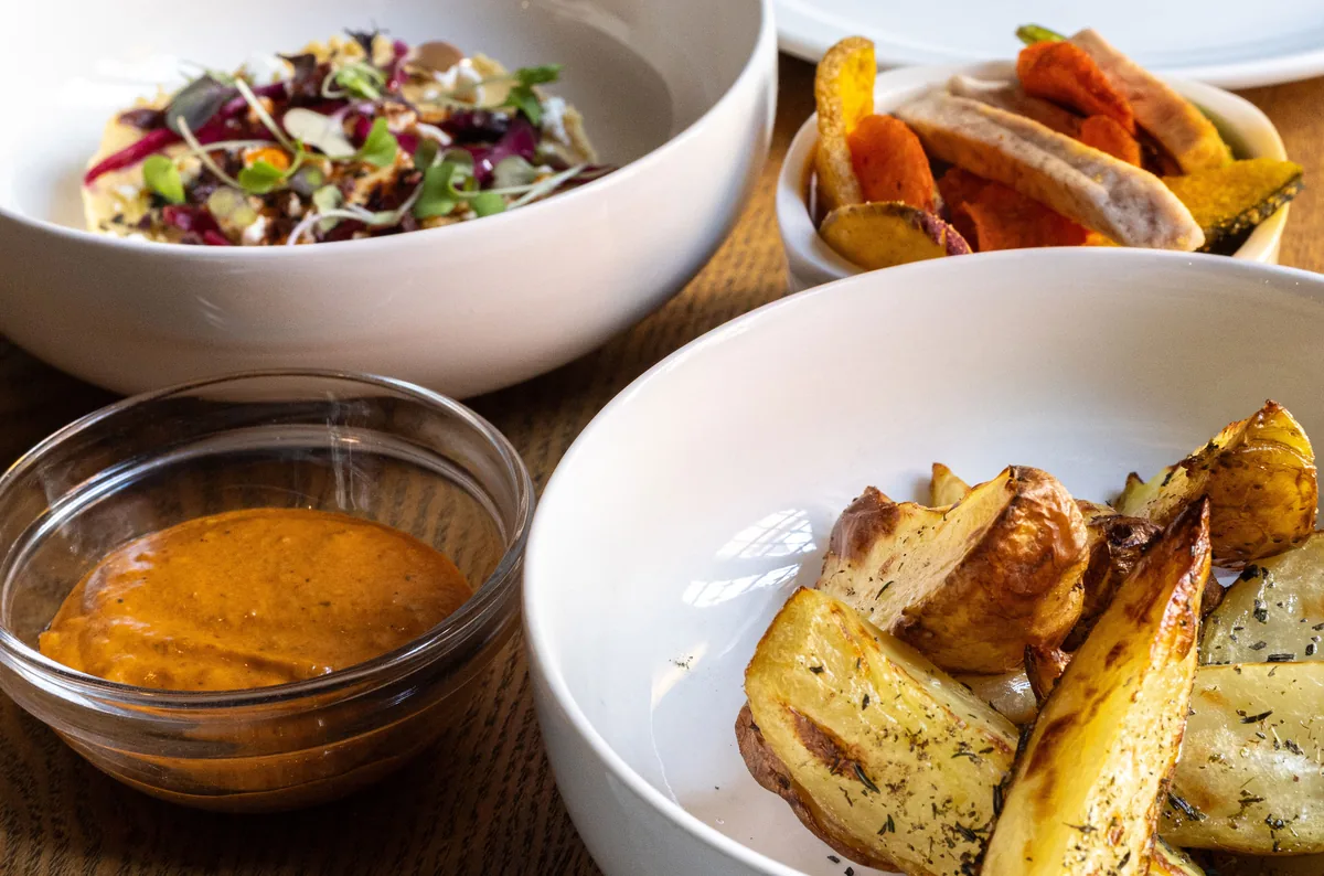 Bowls of salad, herb-roasted potatoes and grilled vegetables with a glass dish of romesco