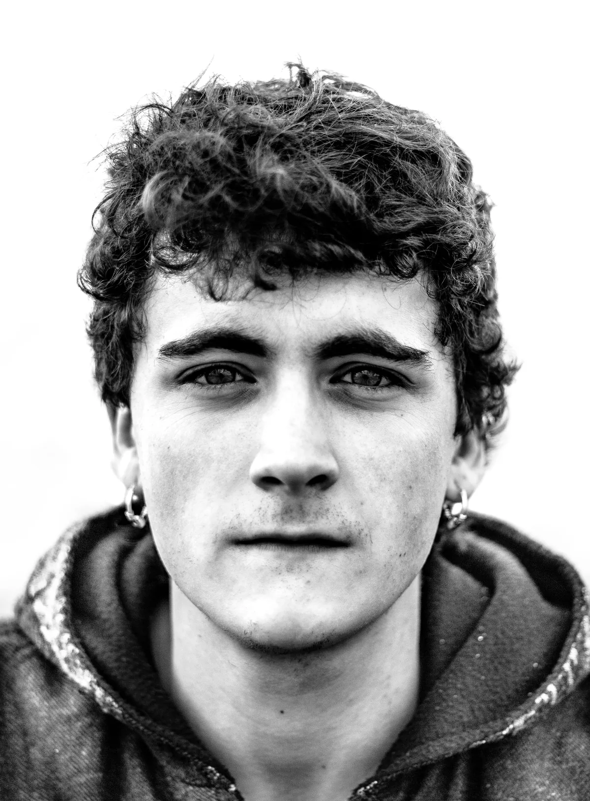 Young man with dark curly hair and hooped earrings in a hooded sweatshirt, looking straight into the camera