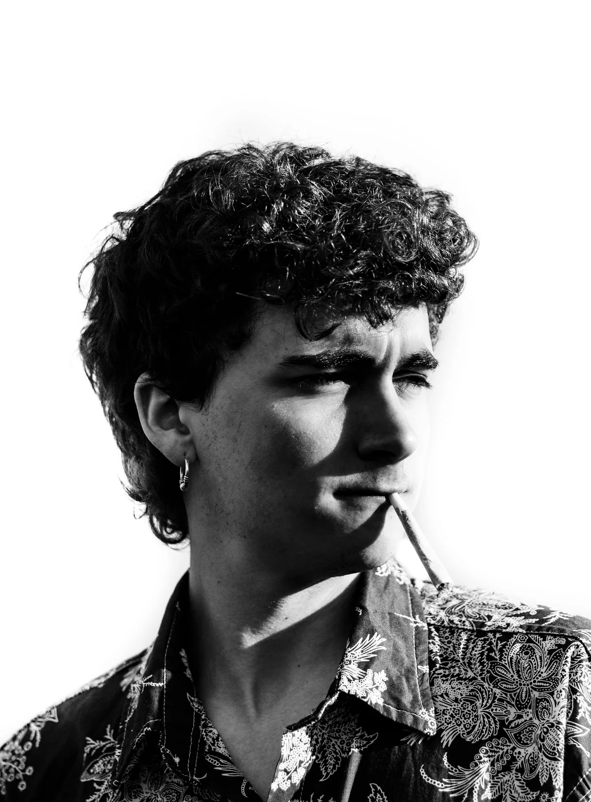 Young man in a patterned shirt with a cigarette between his lips, lit hard against a white background