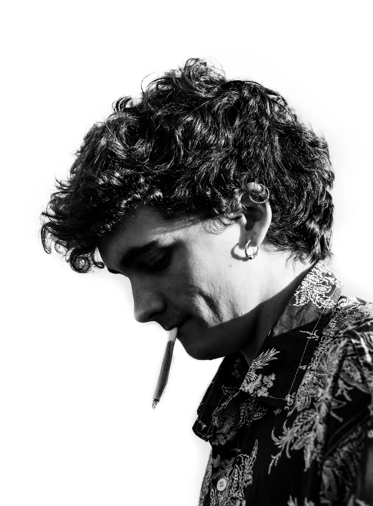 Young man in a patterned shirt photographed from the side, head lowered, cigarette in his mouth