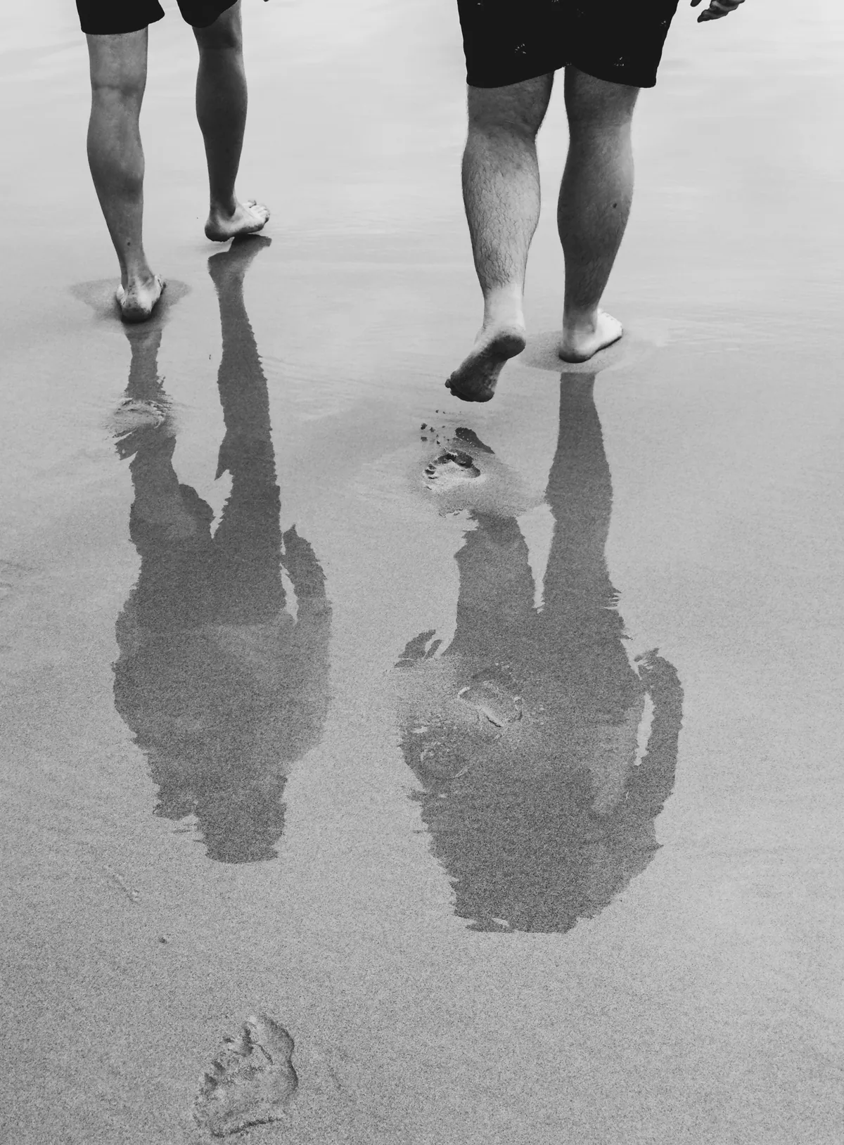 Two people walking barefoot on wet sand, their reflections stretching towards the camera