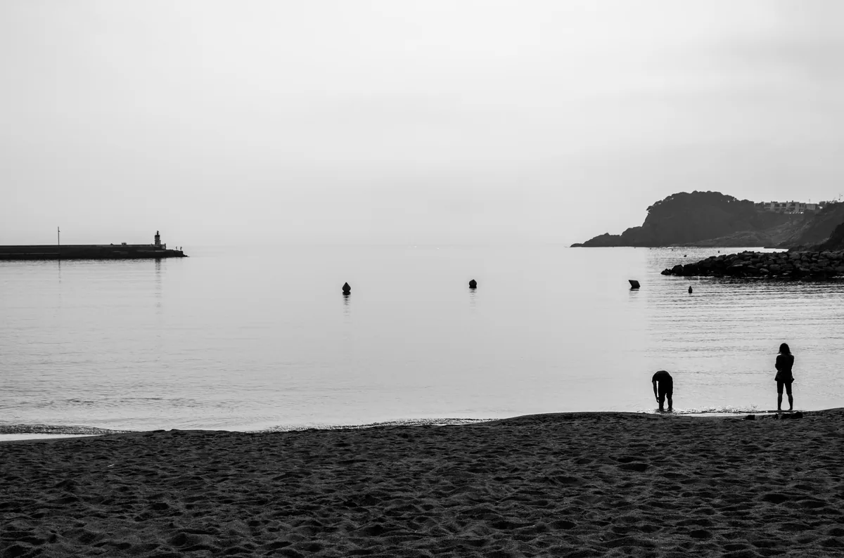 Figures on a beach at dusk, a breakwater and lighthouse on one side and a headland on the other