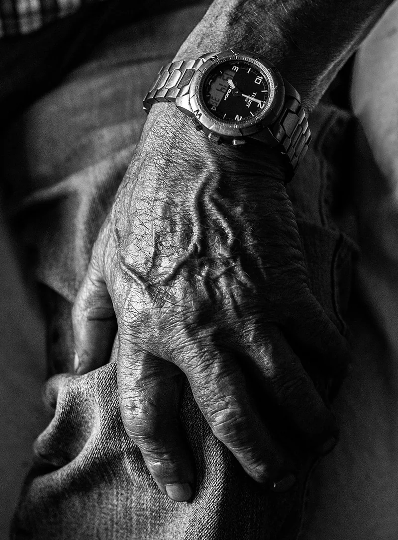 Close-up of an older person's weathered hand and steel wristwatch resting on their knee