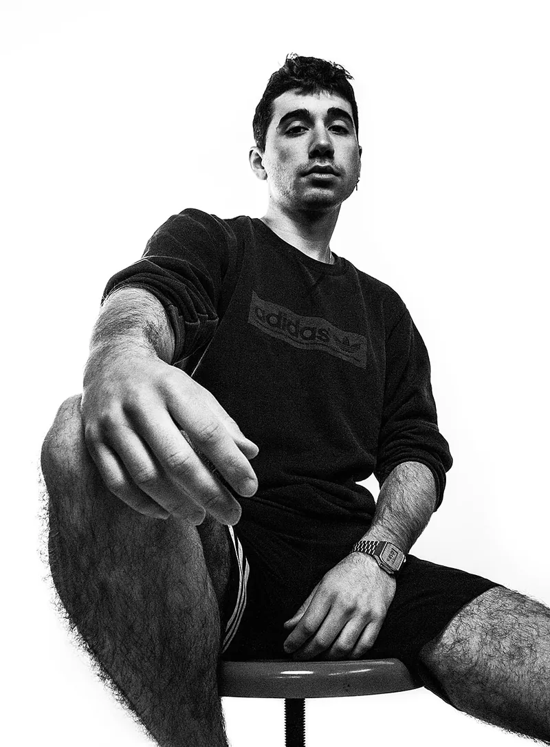 Young man sitting on a stool with one arm reaching towards the lens, shot against white