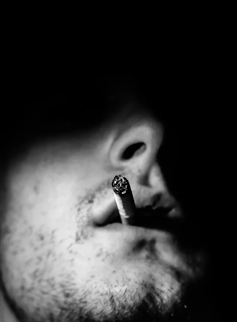 Close-up of a lit cigarette at a man's lips, the rest of the frame in darkness