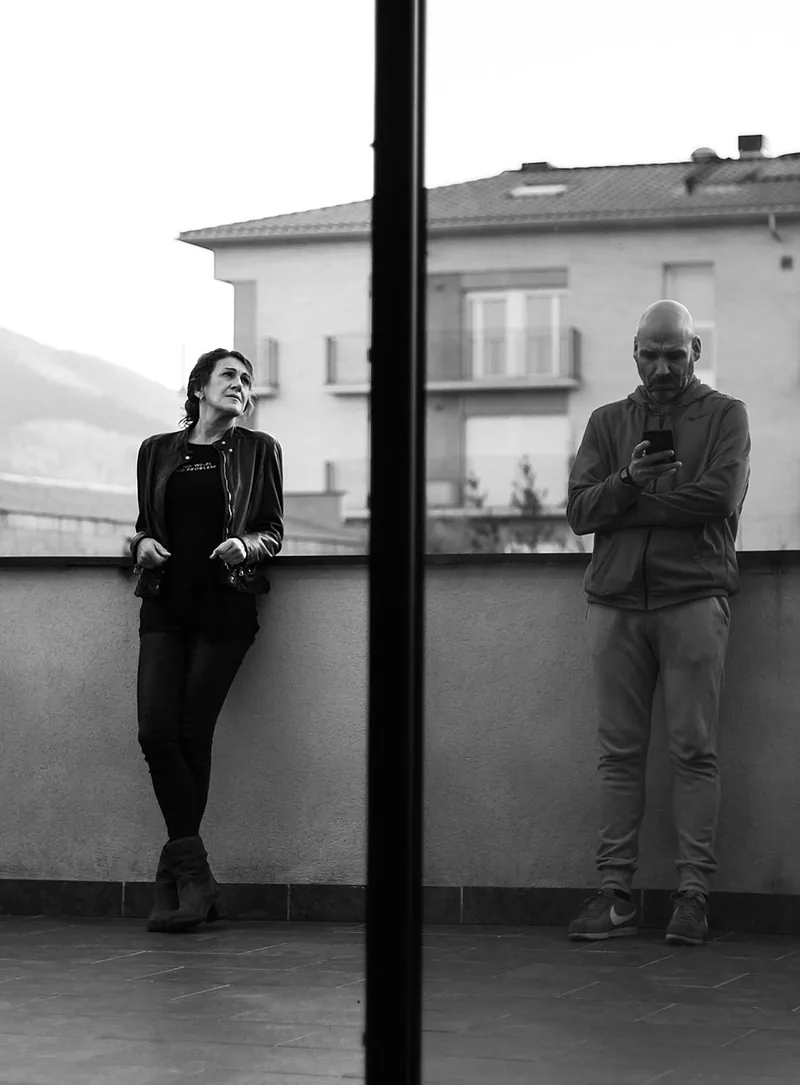 A woman leaning on a balcony rail and a man looking at his phone, seen through a window divided by a dark post