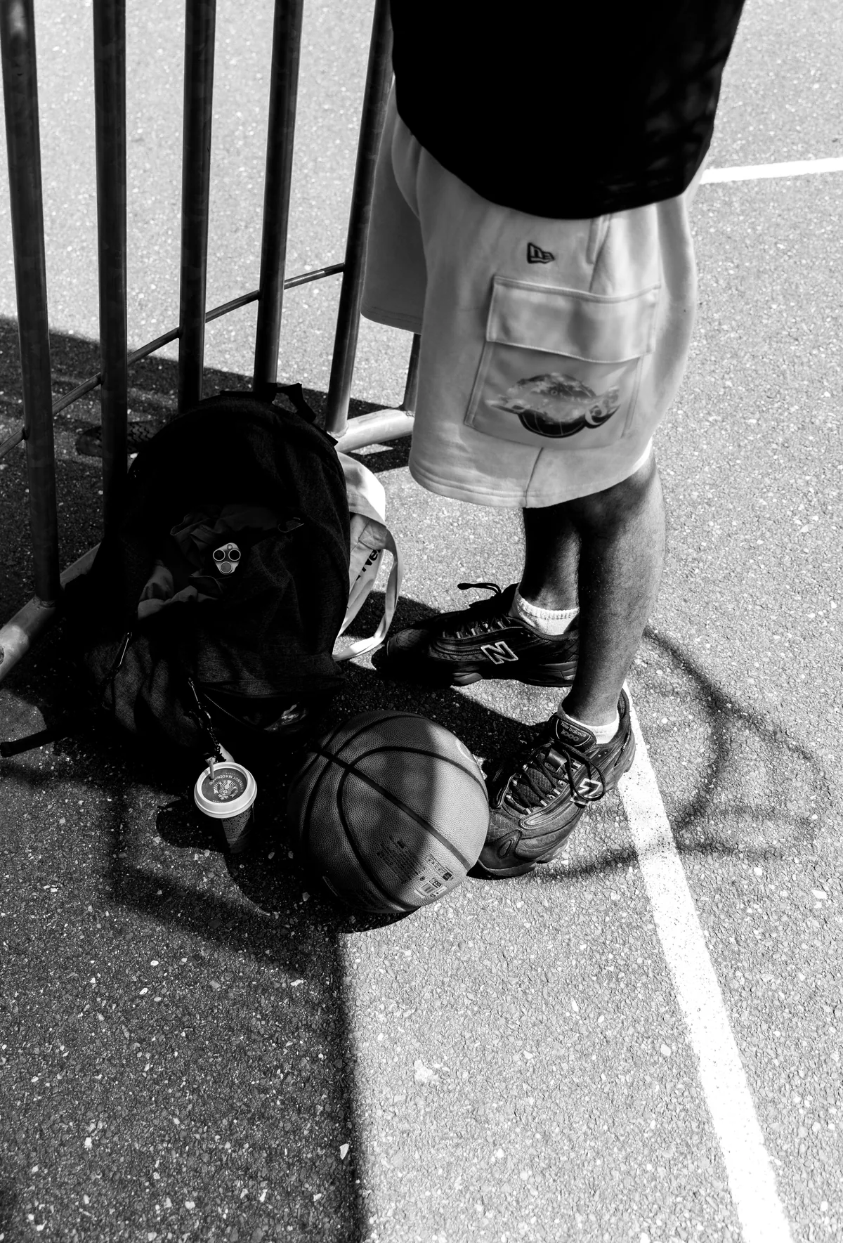 Basketball court at street level: a player's legs, a bag, a ball and a coffee cup by the fence
