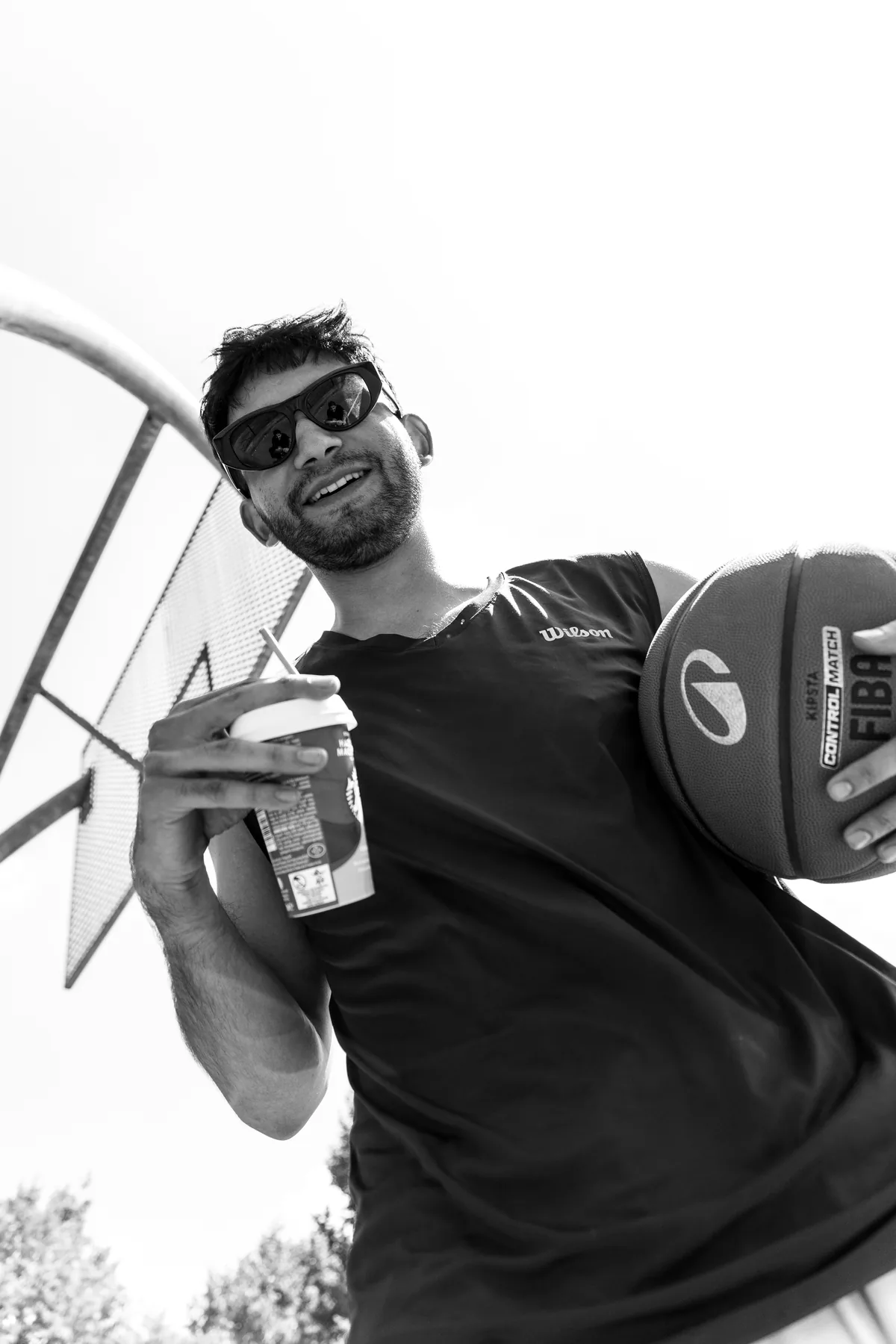 Man in sunglasses holding a basketball and a takeaway coffee, photographed from below against the sky